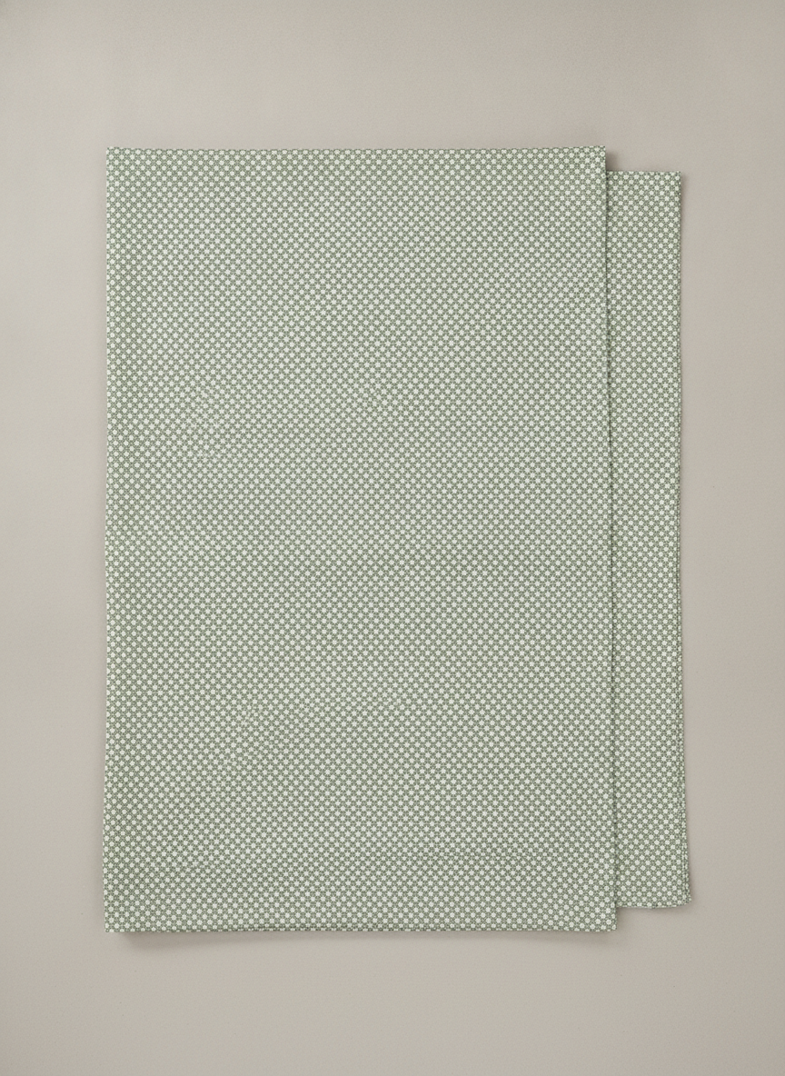 The fabrication view, and the most practically useful picture here: one piece of curtain fabric laid flat and face up on a neutral surface, folded back at one edge so the reverse of the weave shows. A curtain maker can work from this. The full repeat is visible, the density of the small sage and cream geometric is readable, and the design is shown as the design rather than as a lit, gathered, backlit interpretation of it. It is also missing the one thing a swatch must carry. There is no scale reference anywhere in the frame, so a five centimetre repeat and a fifty centimetre repeat produce an identical photograph, and only one of them still reads as a pattern on a three metre drop.
