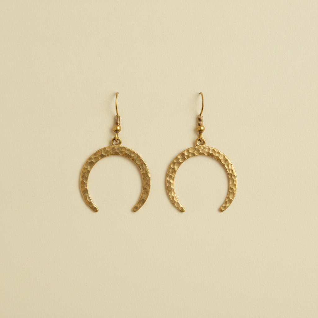 AI Etsy listing image generator result: a pair of hand hammered brass crescent earrings hanging side by side, dead centre, against a seamless warm ivory paper background with nothing else in the square frame. Everything about the craft of this frame is right. The hammered dimpling reads as real planished metal rather than as a texture map, the two earrings genuinely mirror each other, they hang at exactly the same height, the brass is a warm satin rather than a plastic gold, and the frame is completely clean with no prop, no bust, no stand and not one letter anywhere. The single failure is the one number in the brief. The pair was specified to fill about eighty percent of the frame height and came back filling roughly forty, marooned in a wide field of empty ivory. On a marketplace where your first photo is shown as a small square tile next to sixty other small square tiles, that is the whole ballgame, and it is a ten second crop you must remember to do every time.