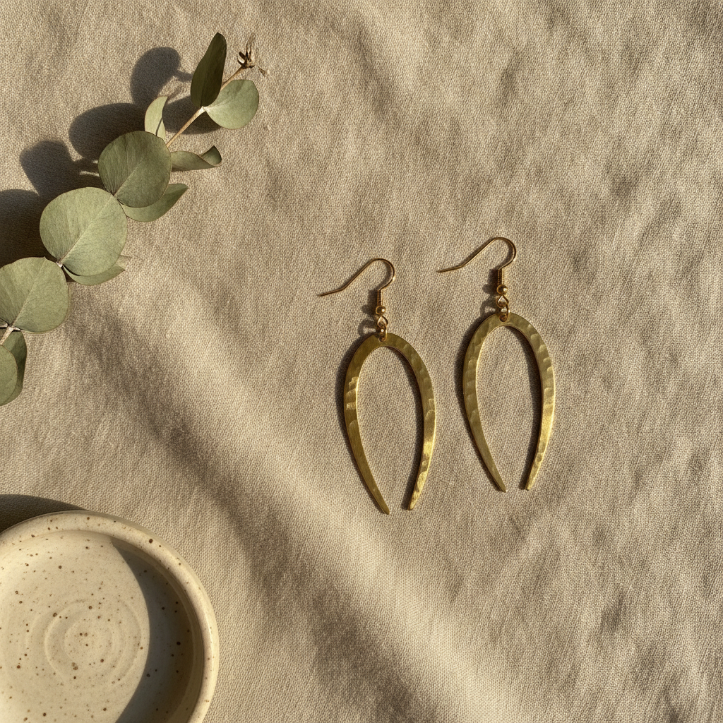 AI Etsy listing image generator result: an overhead flat lay of the brass earrings lying on rumpled oatmeal linen, a dried eucalyptus sprig running across the upper left and a small speckled cream stoneware dish in the lower left, lit by warm low afternoon sun that throws long soft shadows across the cloth. This is the frame you would actually put second in a listing. The light is genuinely raking and directional, the linen creases read as woven cloth rather than as noise, the eucalyptus leaves have the dusty sage bloom that dried eucalyptus really has, and the prop discipline is perfect: exactly the two props asked for arrived and nothing else crept into the frame. Then look at the product. The wide open crescent from the first photo has become a long narrow wishbone, taller, thinner and pointed, with the hammering flattened to almost nothing. Frame one and frame two are already two different earrings.