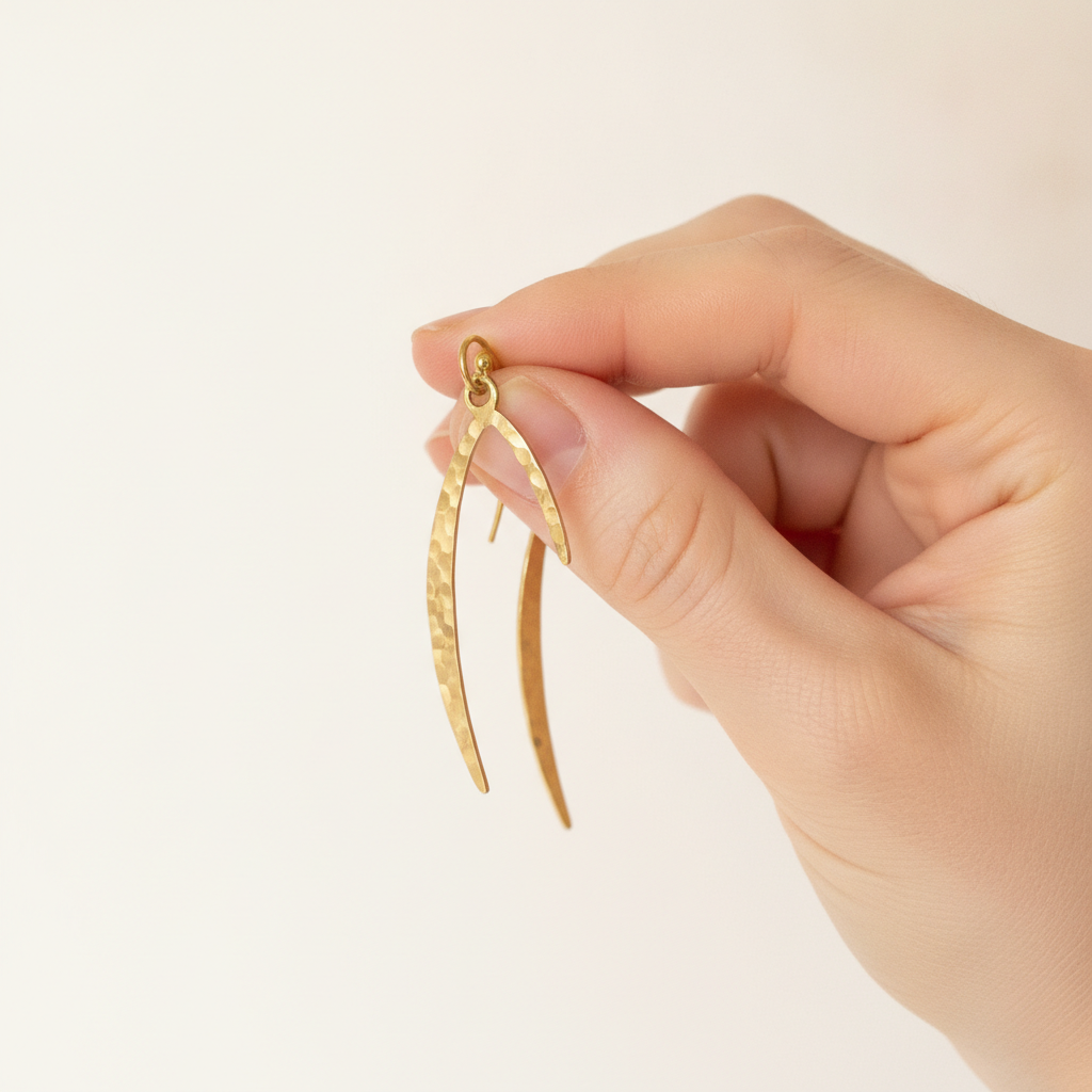 AI Etsy listing image generator result: a scale reference shot with a hand pinching one earring between thumb and forefinger against a plain warm off white background, the second earring hanging just behind it and slightly out of focus. The hand is the part everyone expects to fail and it did not: correct anatomy, believable knuckles and nail beds, a natural pinch grip, no melted sixth finger, no jewellery, no sleeve, and it gives an immediate honest sense of how small the piece is. The background is clean and the brief held on everything it banned, with no ruler, no coin, no tape measure and no lettering anywhere. What it lost was the product again, and this time completely. The closed crescent has become two separate thin curved slivers, the ear wire has vanished behind the fingers on the front earring, and the shape now shares nothing with the first frame except the colour of the metal.