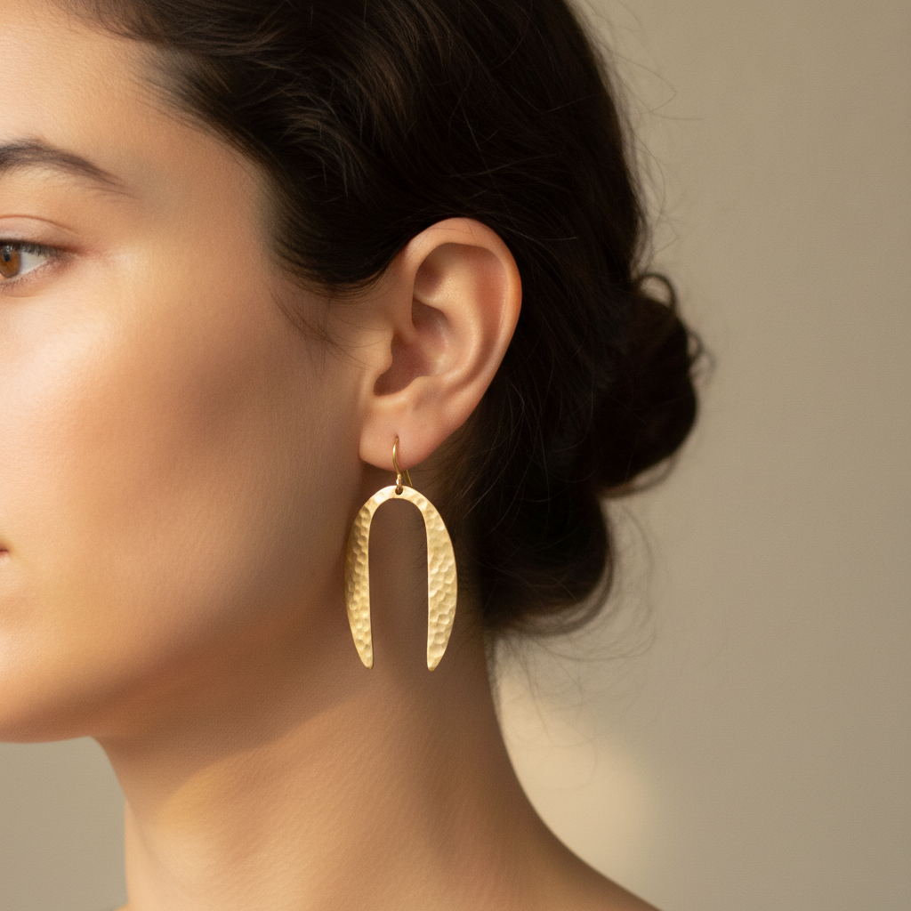 AI Etsy listing image generator result: a worn shot cropped to the side of a head, showing one brass earring hanging from the ear against dark hair pulled back into a low bun, with a soft blurred greige wall behind. As a photograph it is the strongest frame on the page. The window light is soft and directional, the skin has real pores and fine texture instead of the airbrushed plastic this kind of model usually returns, the hair has flyaway strands that catch the light, and the earring sits and hangs the way real metal hangs. Two things are wrong and one of them matters legally rather than aesthetically. The brief said explicitly to turn the face away so that no eyes, no nose and no mouth were visible, and it returned a clear three quarter portrait of an identifiable looking person anyway. And the earring has changed again, into a wide flat inverted horseshoe with the points hanging down, the fourth distinct shape in four frames.