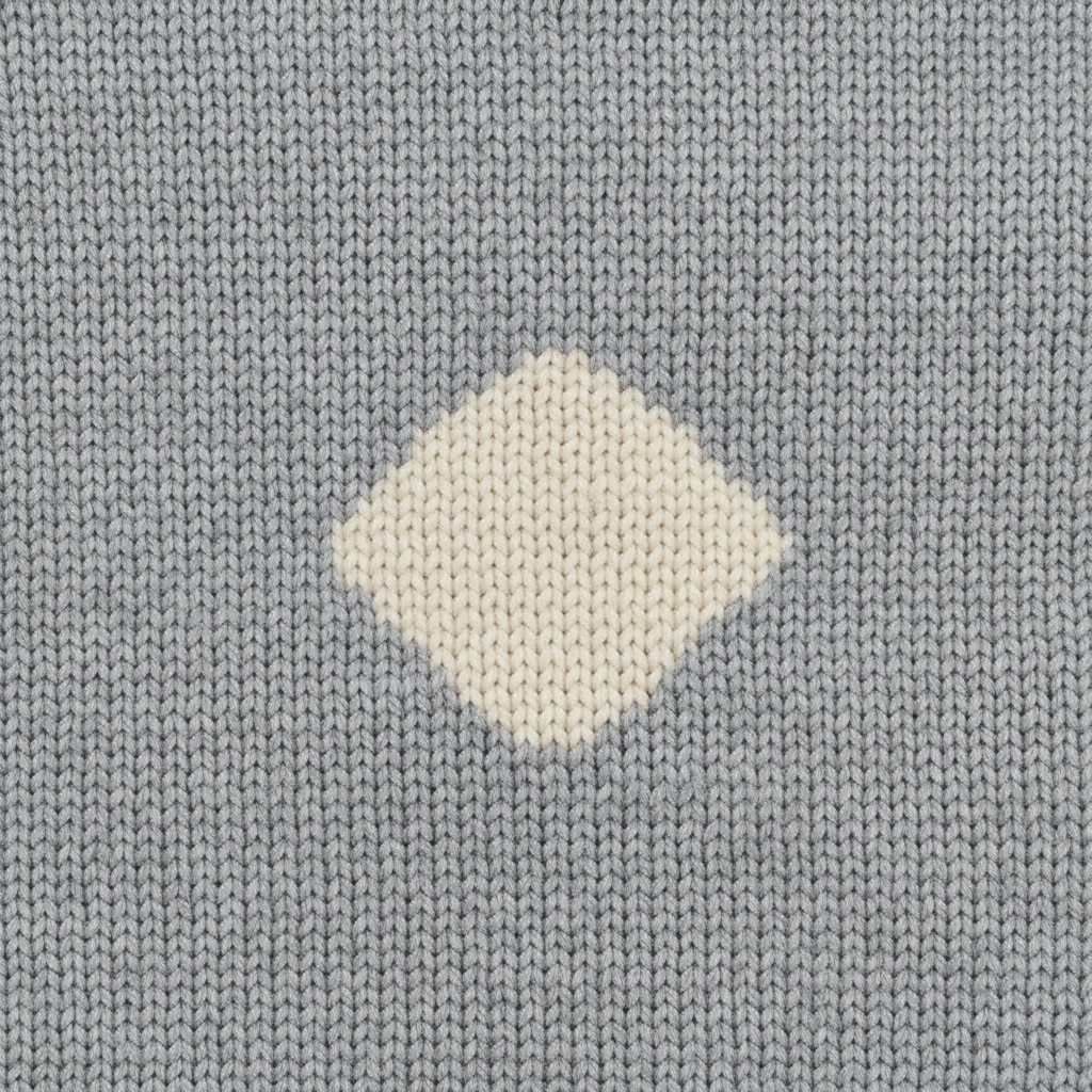 Macro on grey stocking stitch: a cream motif comes out noticeably wider than it is tall because every knitted stitch is a V wider than it is tall, and its edges climb the fabric in visible stair-steps one row at a time