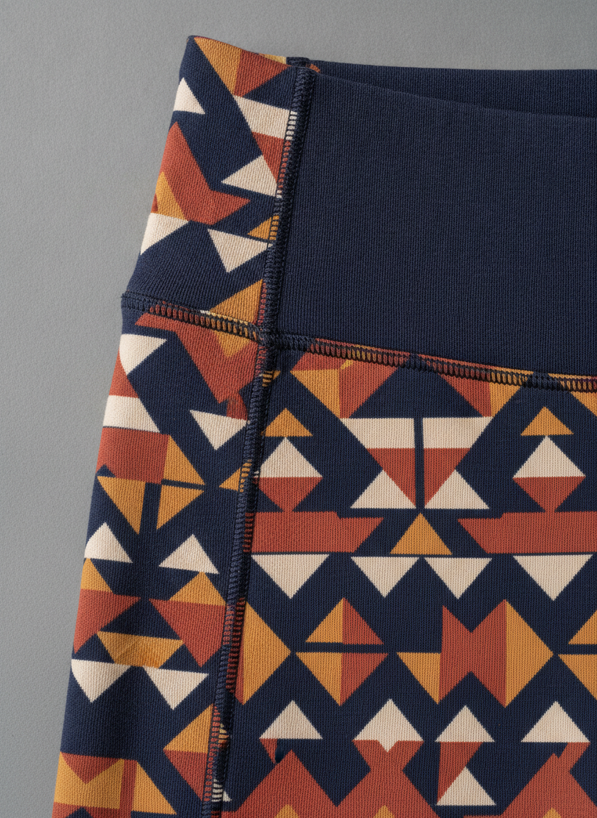 Macro close up of the outside side seam of navy leggings printed with terracotta, mustard and cream triangles, the geometric pattern failing to line up across the flatlock stitching and rendering at a visibly different scale on each side of the seam, with the waistband solid navy on one side of the seam and printed on the other
