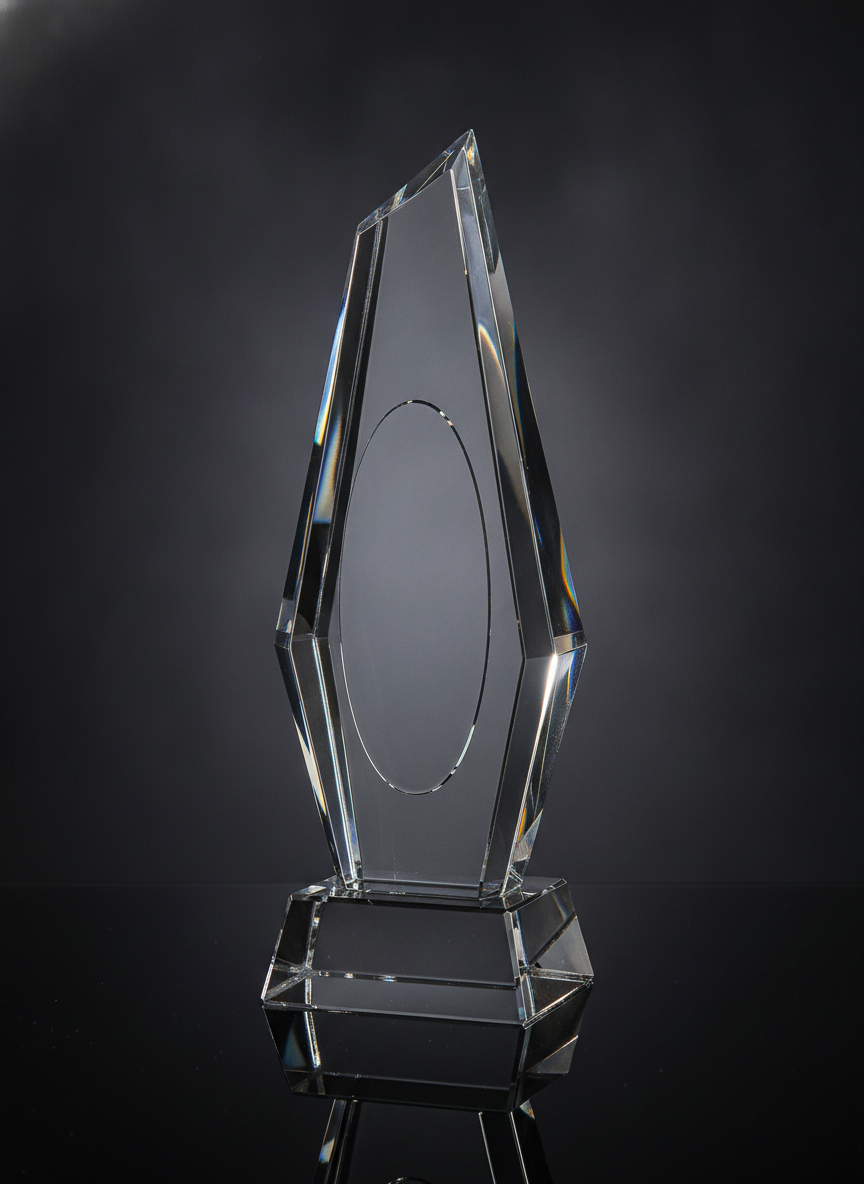 A faceted optical crystal peak on a thick crystal base, photographed against a near-black background and lit from behind so the bevels catch fine spectral colour, with a blank polished oval left in the front face
